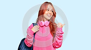 Young redhead woman holding student backpack and books pointing thumb up to the side smiling happy with open mouth