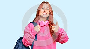 Young redhead woman holding student backpack and books smiling happy and positive, thumb up doing excellent and approval sign