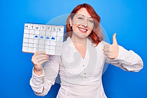 Young redhead woman holding weather calendar showing rainy week over blue background smiling happy and positive, thumb up doing
