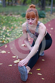 Young redhead woman leg stretching warm up before running on track at autumn