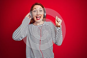 Young redhead woman listening to music using headphones over red isolated background screaming proud and celebrating victory and