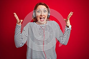 Young redhead woman listening to music using headphones over red isolated background very happy and excited, winner expression