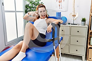 Young redhead woman lying on rehabilitation bed holding crutches pointing fingers to camera with happy and funny face