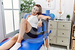 Young redhead woman lying on rehabilitation bed holding crutches sticking tongue out happy with funny expression