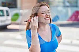 Young redhead woman miling confident listening audio message by the smartphone at street