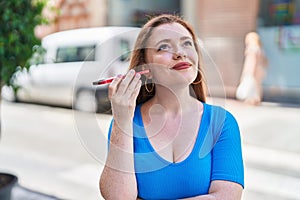 Young redhead woman miling confident listening audio message by the smartphone at street