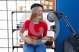 Young redhead woman musician singing song playing ukulele at music studio