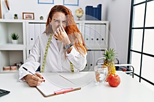 Young redhead woman nutritionist doctor at the clinic smelling something stinky and disgusting, intolerable smell, holding breath