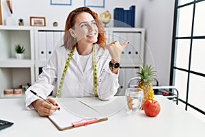 Young redhead woman nutritionist doctor at the clinic smiling with happy face looking and pointing to the side with thumb up