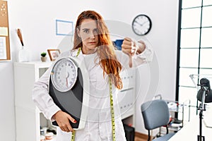 Young redhead woman nutritionist doctor holding weighing machine with angry face, negative sign showing dislike with thumbs down,