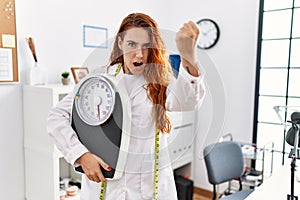 Young redhead woman nutritionist doctor holding weighing machine annoyed and frustrated shouting with anger, yelling crazy with