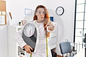 Young redhead woman nutritionist doctor holding weighing machine pointing with finger to the camera and to you, confident gesture
