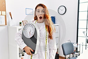 Young redhead woman nutritionist doctor holding weighing machine scared and amazed with open mouth for surprise, disbelief face