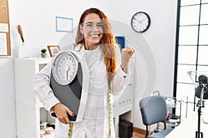 Young redhead woman nutritionist doctor holding weighing machine screaming proud, celebrating victory and success very excited