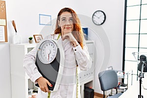 Young redhead woman nutritionist doctor holding weighing machine serious face thinking about question with hand on chin,