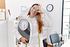 Young redhead woman nutritionist doctor holding weighing machine smiling happy doing ok sign with hand on eye looking through
