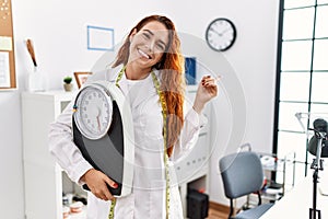 Young redhead woman nutritionist doctor holding weighing machine smiling happy pointing with hand and finger to the side