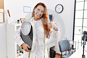 Young redhead woman nutritionist doctor holding weighing machine smiling happy and positive, thumb up doing excellent and approval