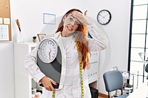 Young redhead woman nutritionist doctor holding weighing machine stressed and frustrated with hand on head, surprised and angry