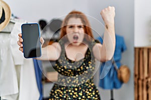 Young redhead woman at retail shop showing smartphone screen annoyed and frustrated shouting with anger, yelling crazy with anger