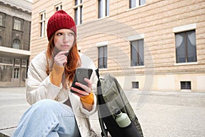 Young redhead woman with smartphone, sitting outdoors with backpack, thinking, looking thoughtful, making decision
