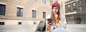 Young redhead woman with smartphone, sitting outdoors with backpack, thinking, looking thoughtful, making decision