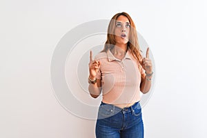 Young redhead woman stading over white isolated background amazed and surprised looking up and pointing with fingers and raised