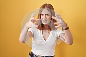 Young redhead woman standing over yellow background shouting frustrated with rage, hands trying to strangle, yelling mad