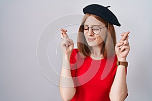 Young redhead woman standing wearing glasses and beret gesturing finger crossed smiling with hope and eyes closed