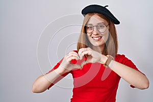 Young redhead woman standing wearing glasses and beret smiling in love doing heart symbol shape with hands
