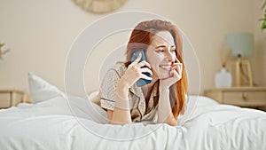 Young redhead woman talking on smartphone lying on bed at bedroom