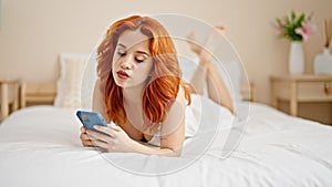 Young redhead woman using smartphone lying on bed at bedroom