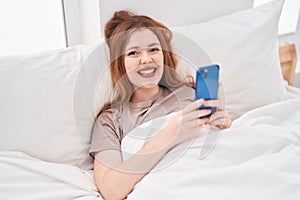 Young redhead woman using smartphone lying on bed at bedroom