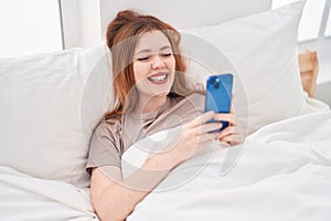 Young redhead woman using smartphone lying on bed at bedroom
