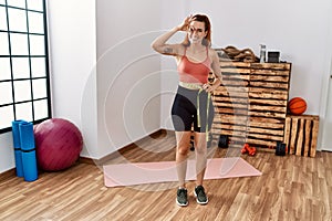 Young redhead woman using tape measure measuring waist at the gym smiling happy doing ok sign with hand on eye looking through