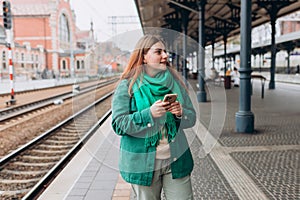 Young redhead woman waiting train with backpack and using smart phone. Railroad transport concept, Traveler. 30s Woman