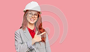 Young redhead woman wearing architect hardhat cheerful with a smile of face pointing with hand and finger up to the side with