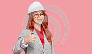 Young redhead woman wearing architect hardhat doing happy thumbs up gesture with hand