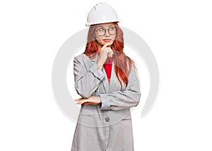 Young redhead woman wearing architect hardhat with hand on chin thinking about question, pensive expression