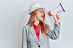 Young redhead woman wearing architect hardhat and megaphone angry and mad screaming frustrated and furious, shouting with anger