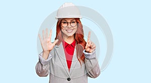 Young redhead woman wearing architect hardhat showing and pointing up with fingers number six while smiling confident and happy