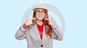Young redhead woman wearing architect hardhat smiling cheerful showing and pointing with fingers teeth and mouth