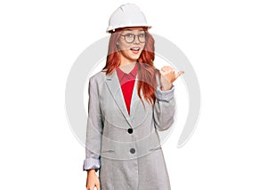 Young redhead woman wearing architect hardhat smiling with happy face looking and pointing to the side with thumb up