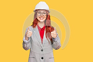 Young redhead woman wearing architect hardhat success sign doing positive gesture with hand, thumbs up smiling and happy