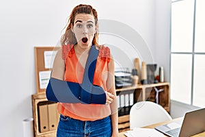 Young redhead woman wearing arm on sling at the office scared and amazed with open mouth for surprise, disbelief face