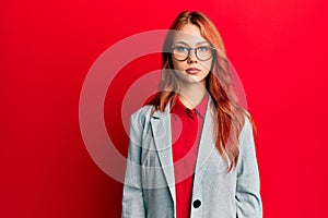Young redhead woman wearing business jacket and glasses with serious expression on face