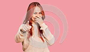 Young redhead woman wearing casual winter sweater laughing at you, pointing finger to the camera with hand over mouth, shame