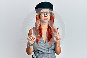 Young redhead woman wearing fashion french look with beret pointing up looking sad and upset, indicating direction with fingers,