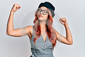 Young redhead woman wearing fashion french look with beret showing arms muscles smiling proud