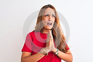 Young redhead woman wearing red casual t-shirt stading over white isolated background begging and praying with hands together with